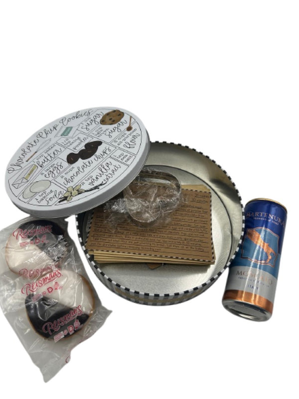 An open Purim gift tin featuring a chocolate chip cookie recipe design on the lid. Inside, the set includes a metal cookie cutter, recipe cards, two Reisman’s Black and White Cookies, and a 375ml can of Bartenura Moscato, creating a charming baking-themed Shalach Manos gift.