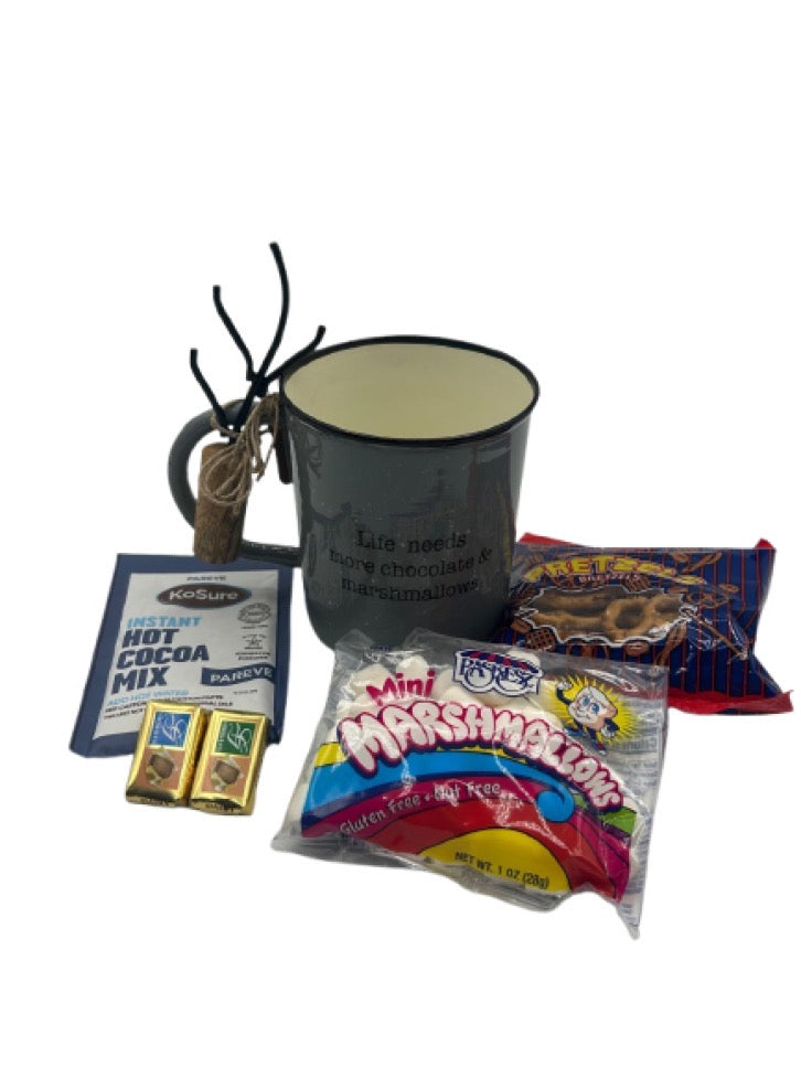 A cozy hot chocolate-themed Shalach Manos gift set featuring a dark gray enamel mug with ‘Life needs more chocolate & marshmallows’ printed on it. The set includes a pack of Ko-Sure Instant Hot Cocoa Mix, a bag of Paskesz Mini Marshmallows, a bag of Lieber’s Pretzels, and two Alprose Chocolate Napolitains. A rustic roasting fork is tied to the mug handle with twine.