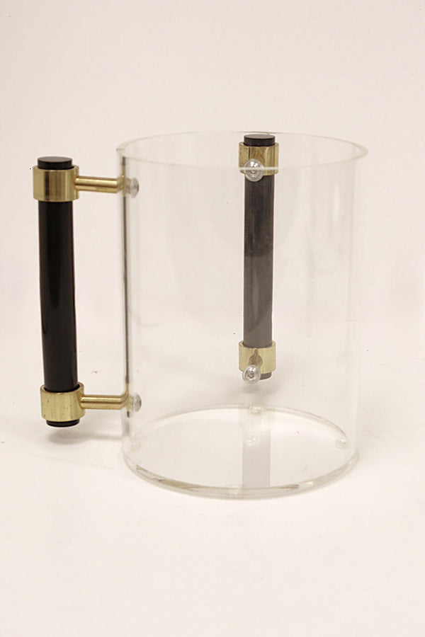 A transparent cylindrical container with a black and gold handle attached to the side. Inside the container, there is a vertical black rod secured by gold fittings at the top and bottom. The container appears to be a stylish modern pitcher or measuring vessel with a minimalist design.