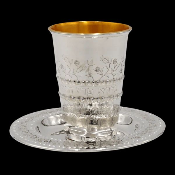  A silver-plated Kiddush cup with intricate floral engravings and Hebrew text, featuring a gold-lined interior, placed on a matching decorative silver plate with cutout designs.