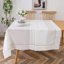 A white tablecloth with silver trim (70" x 120") draped over a wooden dining table, styled in a modern dining room.