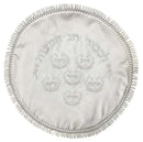 A round, white satin matzah cover with intricate embroidery. It has silver threads showing Hebrew text and six small circles with more Hebrew words. The edge is decorated with a ruffled silver trim.