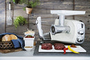 Ankarsrum meat grinder attachment in white, connected to the side of a mixer, with a large white feed tray on top and a silver grinding head. Raw meat is visible on the surface below.