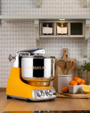 Premium Ankarsrum Original kitchen machine with a vibrant orange base and large, polished stainless steel mixing bowl, ideal for baking and food preparation. This durable stand mixer features chrome accents and a front control panel with two dials for easy operation.