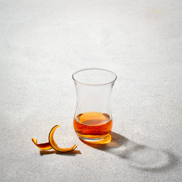 A LAV 4oz Turkish tea glass with a traditional curved shape, partially filled with amber-colored tea, set against a neutral background with an orange peel garnish.