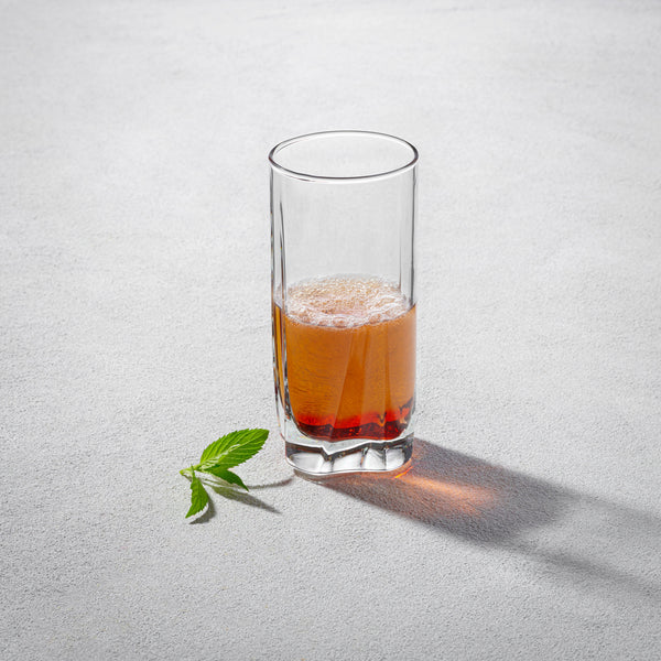 A Vikko Shine 12-ounce clear drinking glass, partially filled with a golden beverage, set on a neutral surface with a fresh green leaf beside it.