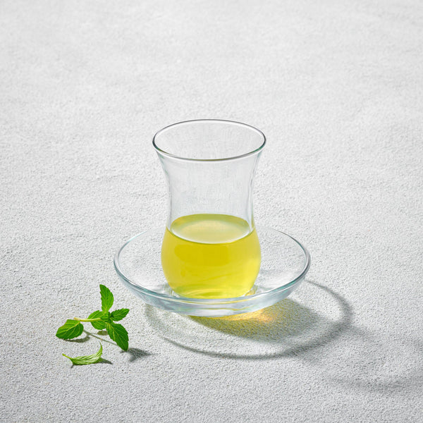 A LAV Turkish tea glass with a curved design, accompanied by a matching clear glass saucer, elegantly displayed with herbal tea.