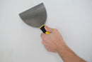 Hand holding a DDON Pro 6-inch flex joint knife against a wall, demonstrating its use for drywall or patching work.