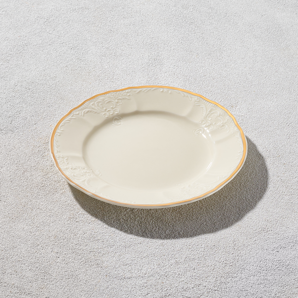 Ivory bread and butter plate with ornate embossed rim and gold edge placed on a textured surface