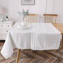 A neatly set table with a white tablecloth, a plate, and a vase of greenery, surrounded by wooden chairs in a bright dining area.