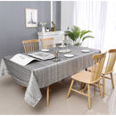 A modern dining table with a patterned tablecloth, set with plates and a candle, surrounded by wooden chairs and a plant.