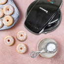 Black Courant donut maker on a countertop with baked donuts and powdered sugar beside it.