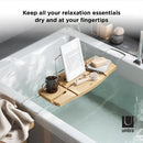 A light wood Umbra bathtub caddy with expandable metal arms, featuring a slotted book/tablet stand, a wine glass holder, and a recessed area with drain holes for soap or accessories.