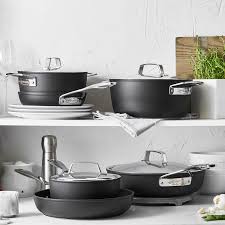 A collection of black non-stick pots and pans with stainless steel handles and glass lids, neatly stacked on a white shelf.