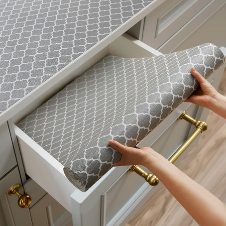 A person placing a roll of gray patterned drawer liner into a kitchen drawer, with the same liner already applied to the countertop above. The liner features a white quatrefoil design and is cut to fit the space.