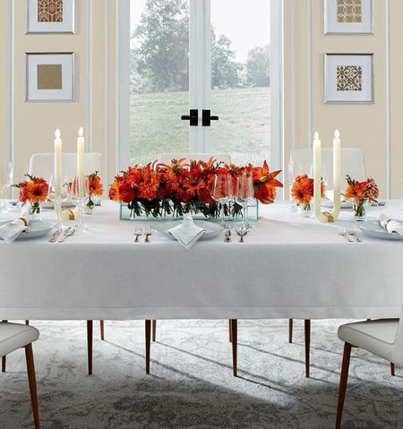 A beautifully set dining table covered with a white tablecloth, adorned with red-orange floral centerpieces, white candles, and fine glassware in a bright dining room with large windows.