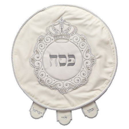 A round white satin Passover matzah cover with intricate silver embroidery, featuring a decorative crown design and the Hebrew word "Pesach" in the center. The cover includes three labeled flaps for holding the three matzot used during the Seder.