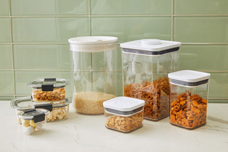 A set of airtight food storage containers of various sizes, made of clear plastic with white lids, filled with dry goods such as rice, pasta, nuts, and pretzels, arranged on a kitchen countertop with a green tile backsplash.
