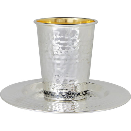A silver-toned hammered Kiddush cup with a gold-plated interior, sitting on a matching round saucer. The elegant design features a textured surface, making it a refined addition to any Shabbat or holiday table.
