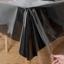 This image shows a clear plastic tablecloth or protector laid over a dark-colored (likely black) tablecloth or table. A hand is visible in the bottom right corner, slightly pulling up an edge of the clear plastic, demonstrating its transparent quality and flexibility. The underlying table appears to be made of wood.