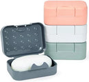 A stack of three colored plastic travel soap dishes (peach, white, and mint green). One dark gray case is open, holding a bar of white soap.