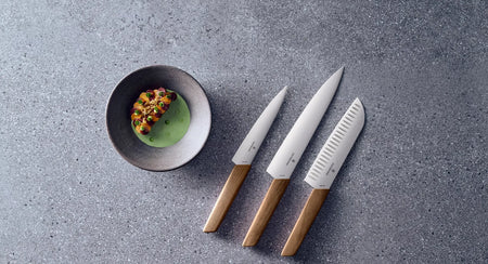 A sleek set of three kitchen knives with wooden handles, arranged on a textured gray surface next to a bowl of food.