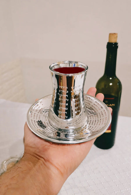 Elegant silver kiddush cup with detailed Hebrew inscriptions and matching base plate, set against a softly lit background.