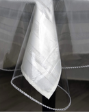 A crystal clear vinyl tablecloth protector from Broder Mfg. Inc. is draped over a white tablecloth with subtle striped patterns. The transparent protector has a delicate white crocheted or lace-like trim along its scalloped edge. The layers create a soft, textured appearance against a dark background, showcasing the protective layer over the fabric tablecloth.