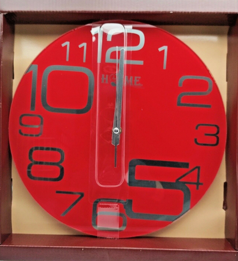 A striking red circular wall clock with large, stylized black numerals, featuring a clear vertical stripe through the center with the brand "HOME" subtly visible. The clock is shown inside an open brown cardboard box.
