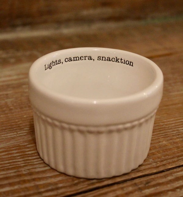A white ceramic ramekin with a fluted exterior and a subtle horizontal beaded band. The black text 'Lights, camera, snacktion' is printed inside the rim.