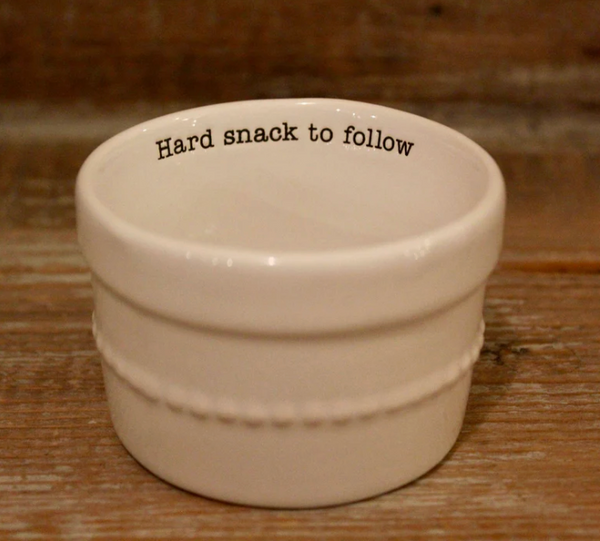 A white ceramic ramekin with a decorative beaded band on the exterior and the black text "Hard snack to follow" printed inside the rim.