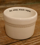 A small, white ceramic ramekin with a subtle stitched texture around the exterior and the black text 'Eat, sleep, snack repeat' printed inside the rim.