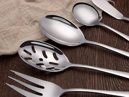 A set of polished stainless steel serving utensils, including spoons, a slotted spoon, a fork, and a knife, laid out on a dark wooden surface with a beige cloth.