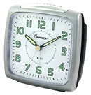 A metallic gray analog alarm clock with a square face, glowing hands, and a light button on top.