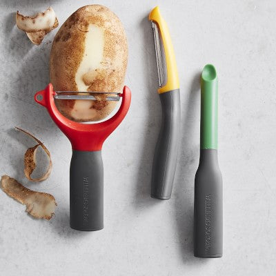 Three colorful peelers with ergonomic handles, including a red Y-peeler holding a partially peeled potato, a yellow straight peeler, and a green corer.