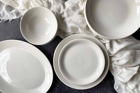 A collection of white ceramic plates and bowls arranged on a dark surface with a soft, cream-colored fabric draped around them.