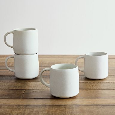 A set of four minimalist white stoneware mugs with a matte finish, placed on a wooden table, with two stacked on top of each other.