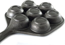 The image displays a cast iron aebleskiver pan. It's a dark-colored pan with a single handle and seven hemispherical indentations, which are used to cook aebleskiver (Danish pancake balls). The shape of the pan, excluding the handle, is hexagonal.