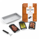 Grilling and serving set by Yukon Glory, featuring a large silver pan and three smaller black grill/sear trays with cooked food, plus a handle, all next to its product packaging.