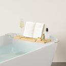 A light wood Umbra bathtub caddy with expandable metal arms, featuring a slotted book/tablet stand, a wine glass holder, and a recessed area with drain holes for soap or accessories.