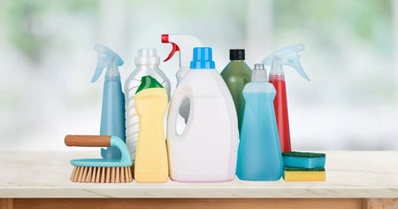 A collection of various cleaning supplies arranged on a wooden countertop, including spray bottles, detergent containers, sponges, and a scrubbing brush. The background is blurred, suggesting a bright and clean indoor environment.