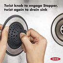 OXO sink accessory with a black center that can function as a strainer or a stopper, surrounded by a stainless steel ring and a gray silicone outer edge.