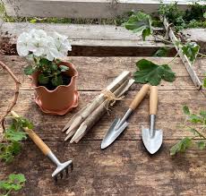 Rustic wooden gardening table with small hand tools, wooden stakes, and a potted white flower surrounded by greenery