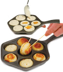 The image displays a cast iron aebleskiver pan. It's a dark-colored pan with a single handle and seven hemispherical indentations, which are used to cook aebleskiver (Danish pancake balls). The shape of the pan, excluding the handle, is hexagonal.