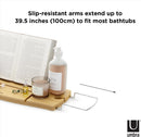 A light wood Umbra bathtub caddy with expandable metal arms, featuring a slotted book/tablet stand, a wine glass holder, and a recessed area with drain holes for soap or accessories.