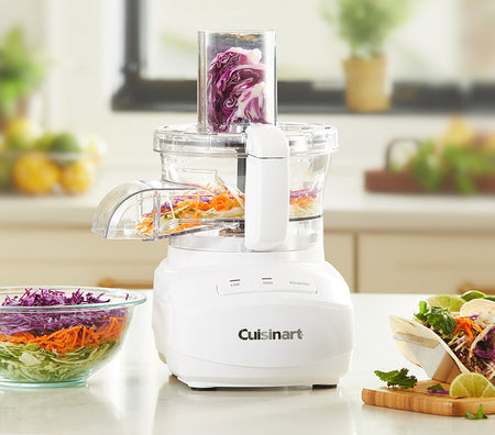A food processor with multiple attachments, chopping vegetables in a clear bowl with a stainless steel base.