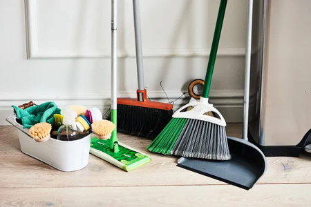 A collection of cleaning tools, including a green mop, two brooms with different colored bristles, a dustpan, and a cleaning caddy filled with brushes, a spray bottle, and microfiber cloths, all placed against a white wall on a wooden floor.
