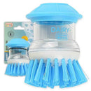 A "Kosher Cook Dairy Blue Sud-Zee Scrub Brush with Liquid Dish Soap Dispenser" is prominently displayed against a plain white background. The brush features bright blue bristles and a clear cylindrical body that holds liquid soap, topped with a blue, mushroom-shaped cap. The word "DAIRY" is visible on the transparent section. In the background, a smaller, similar brush and its packaging are partially visible, reinforcing the product's identity.