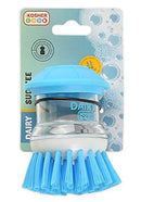 A "Kosher Cook Scrub Brush with Liquid Soap Dispenser" is prominently displayed against a plain background. The brush features a clear, cylindrical body for liquid soap with "KOSHER COOK" and a red "K" logo visible, topped by a light blue, mushroom-shaped cap. Its bright blue bristles flare out from the white base. To the left, part of the packaging is visible, showing the brand "KOSHER COOK" and the "SUD ZEE" product name on a green and blue background with soap bubble graphics.