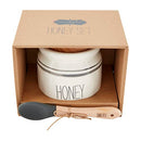 A Mud Pie honey set packaged in a brown box. The set includes a round, white ceramic pot with gray stripes and the word 'HONEY', a wooden lid, and a wooden spoon with a gray silicone end.